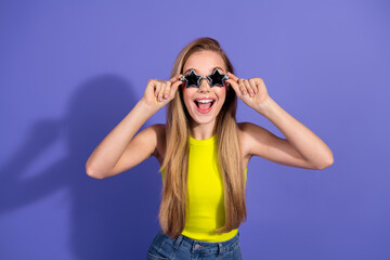 Cheerful young woman with blonde hair in casual summer outfit posing joyfully with star-shaped glasses against a purple background