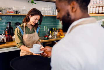 Female barista working in a coffee shop at the counter and gives a coffee to a male waiter to serve to the customer.