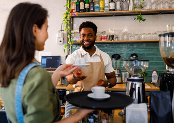 Male barista working in a coffee shop at the counter and gives a coffee to a female waiter to serve to the customer.