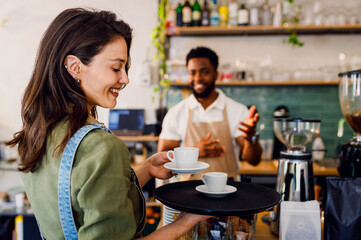 Male barista working in a coffee shop at the counter and gives a coffee to a female waiter to serve to the customer.