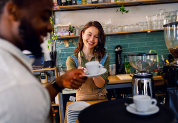 Female barista working in a coffee shop at the counter and gives a coffee to a male waiter to serve to the customer.