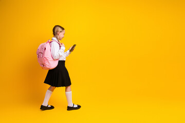Young schoolgirl walking with a backpack and smartphone on a vibrant yellow background, showcasing...
