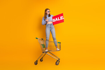 Young woman posing stylishly in a shopping cart holding a sale sign against a yellow background,...