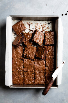 A baking tray filled with freshly cut chocolate brownies, accompanied by a knife on a parchment-lined surface.