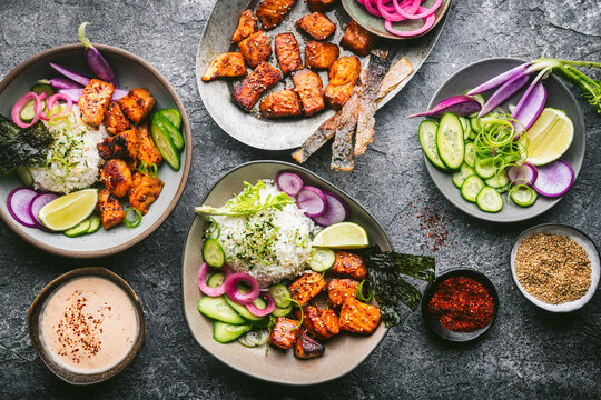 An overhead shot with a custom gray stone textured surface with two beautiful ceramic bowls filled with rice, fresh vegetables, seaweed, and crispy, golden brown pieces of salmon. On the table is a pl