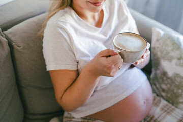 Pregnant woman in pajamas sitting on the sofa and drinking hot cappuccino. Morning coffee.
