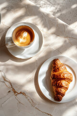 A fresh croissant and a cup of cappuccino with latte art served on a white ceramic table, bathed in morning sunlight with artistic shadows and natural marble texture.