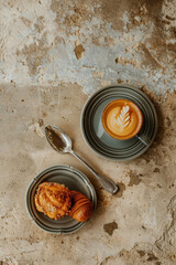 A cup of latte art coffee and two pastries served on elegant gray ceramic plates on a textured stone surface. Overhead composition with a spoon creates a warm, rustic café atmosphere.