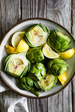 A bowl of fresh artichokes and lemon wedges on a wooden surface.