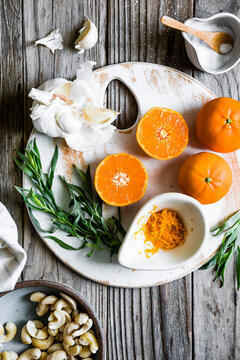 An overhead view of fresh ingredients including oranges, garlic, tarragon, cashews, and salt on a wooden table.