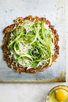 A crispy potato rosti topped with shaved asparagus, grated cheese, and fresh greens on a baking sheet, with a raw egg in a glass beside it.