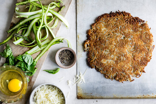 An overhead view of a freshly cooked potato rosti on a baking sheet, with ingredients like egg, cheese, asparagus, basil, and pepper on the side.