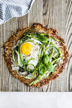 A rustic potato rosti topped with a fried egg, asparagus, and fresh basil on a wooden surface.