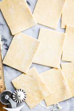 Squares of dough are arranged on a marble surface with a pastry cutter and ruler nearby.