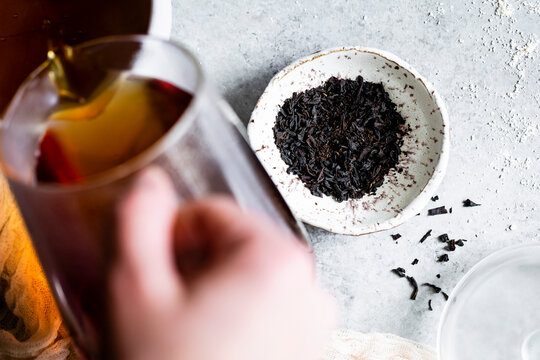 A person pours tea into a glass near a bowl of loose black tea leaves on a textured surface.