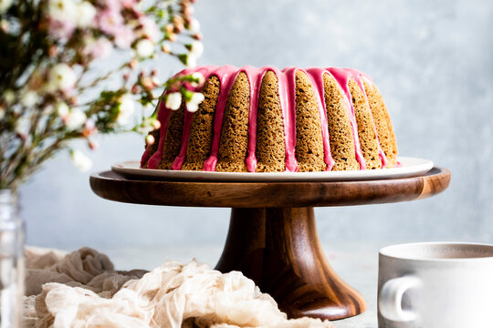 A bundt cake with pink icing is displayed on a wooden cake stand, with flowers and a cup nearby.