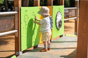 A baby boy wearing a sun hat, striped shirt, and shorts explores a playground. He is interacting with a green activity panel featuring simple drawings designed for sensory play. 