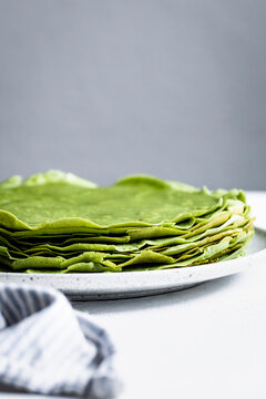 A stack of green crepes on a white plate with a gray background.