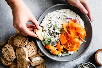 A person prepares to dip a slice of bread into a bowl of creamy dip garnished with carrot ribbons, dill, capers, and red onion, on a textured surface.