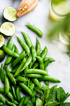 A collection of fresh green sugar snap peas, mint leaves, and lime halves with a wooden citrus reamer on a marble surface.