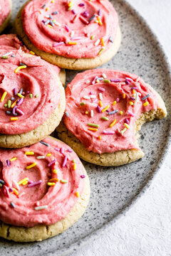 A plate of sugar cookies with pink frosting and colorful sprinkles, one of which has a bite taken out of it.