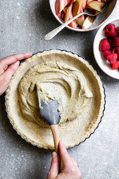 A person spreads filling into a pie crust with a spatula, surrounded by bowls of rhubarb and raspberries.