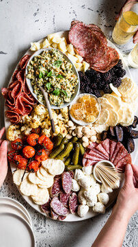 An assorted charcuterie board featuring meats, cheeses, crackers, fruits, nuts, and pickles, with a person holding a glass of champagne.