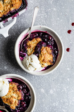 Two bowls of blueberry cobbler with vanilla ice cream on a speckled gray surface.