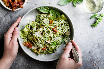 A person holding a bowl of zucchini noodles with pesto, bacon, and parmesan cheese, with a fork ready to eat.