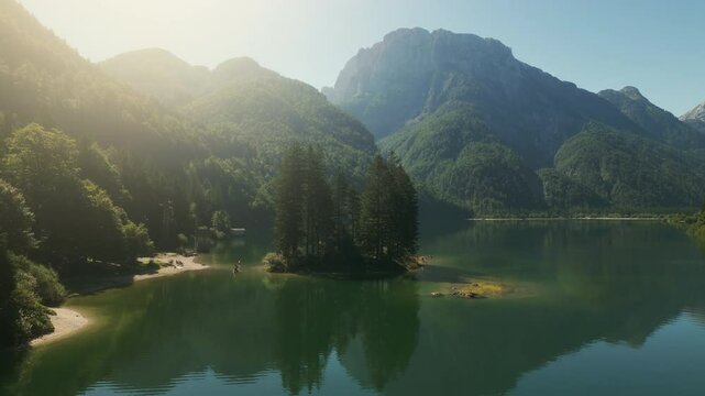 Early morning light bathes the calm Lake Predil surrounded by majestic mountains. Lush trees create an island, reflecting nature's beauty in a tranquil atmosphere.