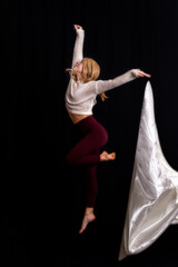 captures a dancer in a graceful pose on a black box against a dark backdrop. Dressed in a light gray sweater and maroon leggings, the dancer extends their arms upward while maintaining balance with on