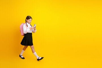 Happy schoolgirl in uniform with pink backpack using smartphone against vibrant yellow background