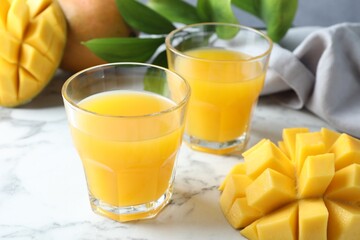 Tasty mango juice in glasses, fresh fruits and green leaves on white marble table, closeup