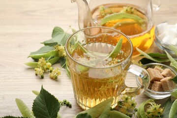 Fresh linden tea in cup, teapot, sugar cubes and flowers on wooden table, closeup