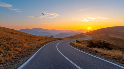 Naklejka premium Curving road leads towards the horizon at sunrise with golden light illuminating the tranquil countryside.