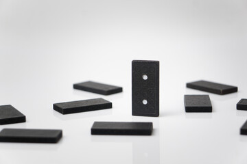 A black domino piece stands upright amidst others lying flat on a reflective white surface.