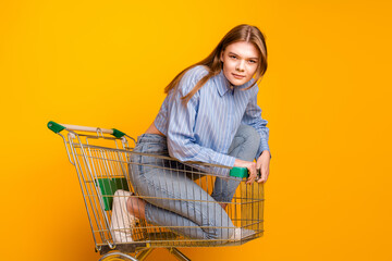 Young woman sitting playfully in a shopping cart on a vibrant yellow background, embodying fun,...