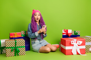Excited young woman with dyed purple hair sits among colorful gift boxes holding a smartphone...