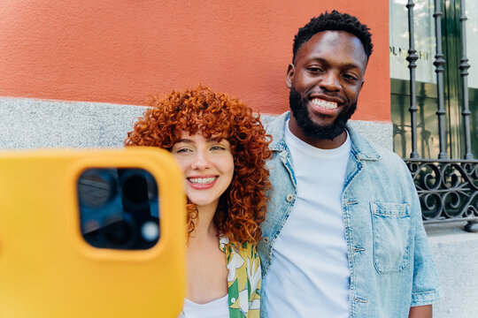 Multiracial couple taking selfie near bright orange wall with metal grating, sharing playful moment in urban environment