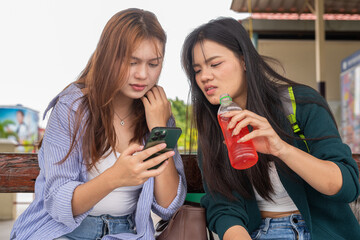 Two young women at train station sitting drinking and using smartphone