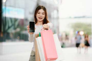 Fototapeta premium A young Asian woman with braces smiles while holding shopping bags and a smartphone outside a store.