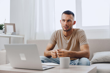 Thoughtful man engaged in a virtual meeting, focusing on the screen while sitting comfortably in a bright living room. Ideal for remote work themes and digital transformation.
