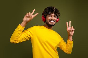 Young man enjoying music in yellow shirt with vivid headphones making peace signs on a green studio background