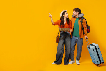 Happy couple preparing for vacation, standing with suitcase and holding map, standing on yellow background and pointing aside at free space