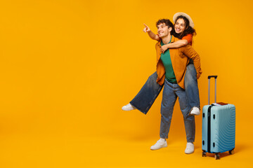 Crazy travel offer. Overjoyed woman piggybacking man, standing with luggage and pointing at free space on yellow background, full length