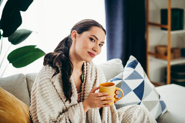 Young woman enjoying leisure time at home, wrapped in a cozy blanket while holding a warm drink in...