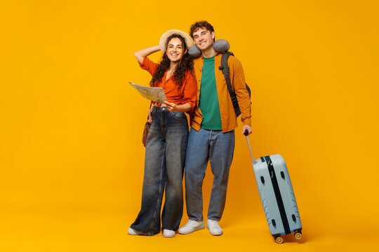 Ready for vacation. Couple holding map, man standing with suitcase, wearing travel neck pillow and backpack, showing their readiness for holiday adventure, yellow background
