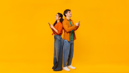 Mobile advertisement. Couple using cellphones, standing back to back on orange studio background, full length, panorama