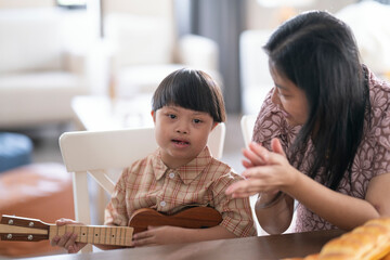 A child with Down syndrome playing ukulele while mother claps along, encouraging musical...