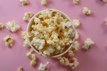 Popcorn in a paper cup. The cup is overflowing and the popcorn is scattered everywhere. Pink background.